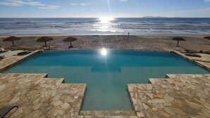 A pool with stone-tiled edges, a beach with umbrellas, and a clear view of the ocean.