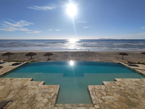 A pool with stone-tiled edges, a beach with umbrellas, and a clear view of the ocean. - Oceanfront 3BR Baja - Pool, Sauna, Gym & Beach (Playas de Rosarito)