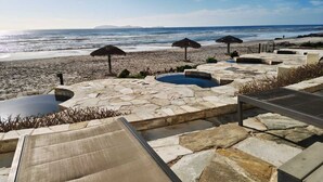 A stone pool area with lounge chairs, a thatched umbrella, and a view of the beach and ocean.