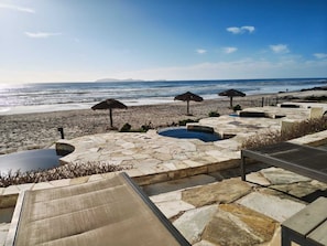 A stone pool area with lounge chairs, a thatched umbrella, and a view of the beach and ocean. - Oceanfront 3BR Baja - Pool, Sauna, Gym & Beach (Playas de Rosarito)