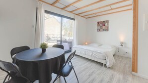 Een kamer met een bed, een tafel met stoelen, een kleine plant in een pot en een raam met gordijnen.