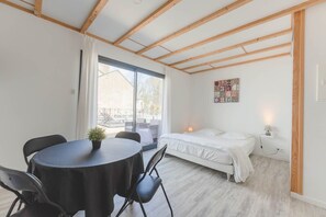 Una habitación con una cama, una mesa con sillas, una planta pequeña en maceta y una ventana con cortinas.