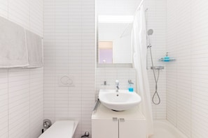 Casa de banho com paredes em azulejos brancos, lavatório branco, espelho, chuveiro e toalha pendurada na parede.