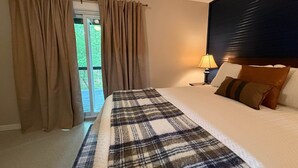 Hotellihuone, jossa on sänky, verhot, yöpöytä, lamppu ja näkymä vihreälle alueelle.
