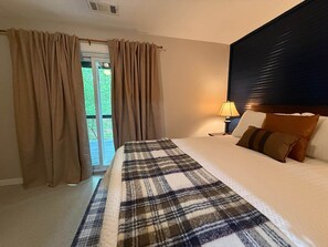 A hotel room with a bed, curtains, a nightstand, a lamp, and a view of greenery outside. - Hot Springs Village Renovated Townhome Retreat (Hot Springs Village)