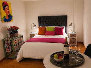 Een netjes opgemaakte slaapkamer met een bed, nachtkastjes, een decoratieve mand, een schaal met fruit, een fles wijn en twee glaasjes.