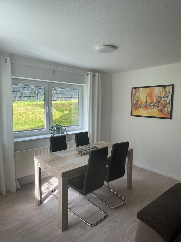 Ein Essbereich mit Tisch, Stühlen, einem Fenster mit Blick auf Grünflächen, einem Heizkörper und einem Bild an der Wand.