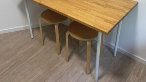 Una mesa rectangular de madera con dos sillas, una pared blanca y un suelo de madera.