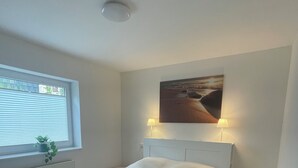 Un dormitorio con cama, ventana con persianas, radiador, un cuadro en la pared y dos lámparas de noche.