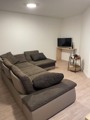 Ein modernes Wohnzimmer mit einem Ecksofa, einem Flachbildfernseher auf einem Holzständer und einem Korbwägelchen auf einem kleinen Tisch.