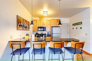 A modern kitchen with wooden cabinets, a central island with bar stools, and a 'HOME AGAIN' sign on the wall. - Ski-In/Out Condo w/ Fireplace + Dogs OK (Whitefish)