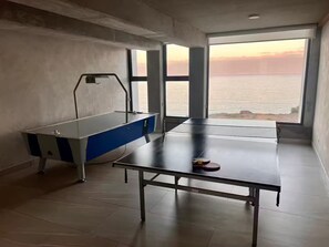 Ein Zimmer mit einem Pongtisch, einem blau-weißen Billardtisch und großen Fenstern mit Meerblick.