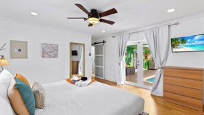Habitación de hotel con cama, ventilador de techo, televisión, puerta corredera a un balcón y vista a la piscina.