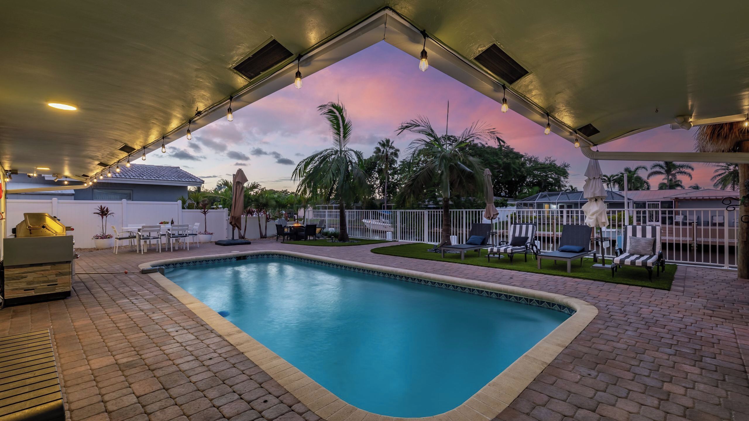 A swimming pool with lounge chairs and umbrellas, surrounded by palm trees and a sunset view.
