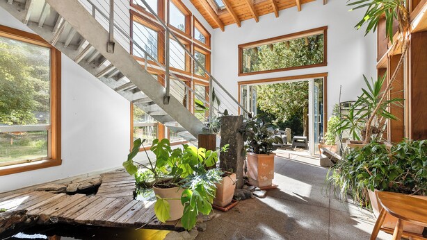 Un interior espacioso con una escalera de madera, ventanas amplias y plantas en macetas.