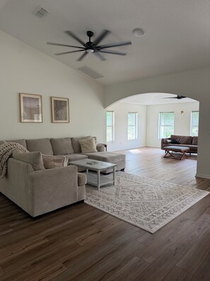 A spacious living room with a large sectional sofa, a coffee table, and a ceiling fan. There are two framed pictures on the wall and a view of the adjacent dining area. - Private 3/2 5 min from WEC Ocala (Ocala)