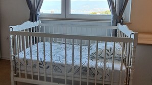 Una habitació amb una bressol blanca, una finestra amb cortines i vista al mar.