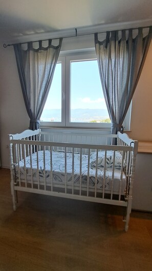 Een kamer met een wit wiegje, een raam met gordijnen en uitzicht op zee.