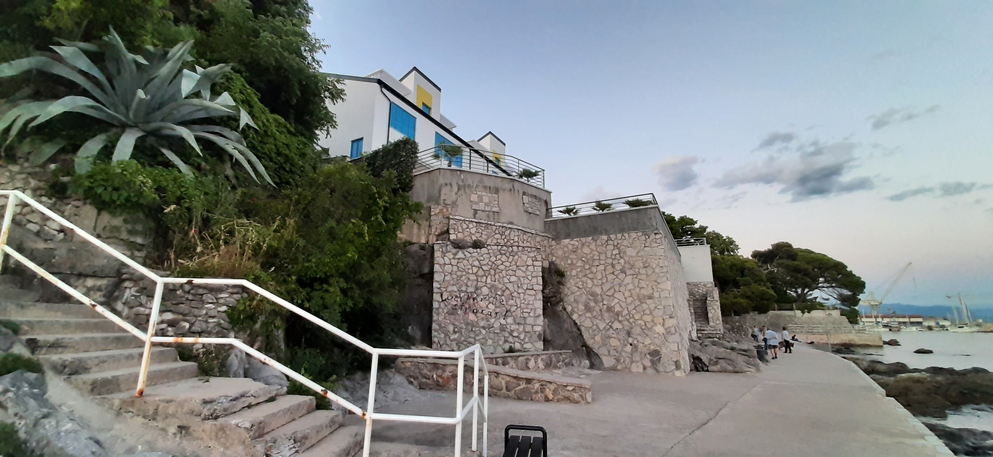 Un edificio costero con escalones de piedra que conducen a un balcón, rodeado de vegetación.
