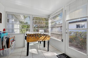 Ein Zimmer mit Kicker, einem Fenster mit Jalousien, einer Tür und Blick auf Grünfläche.