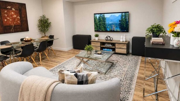 A modern living room with a glass coffee table, a dining area with a glass table, black chairs, and a TV mounted on the wall.
