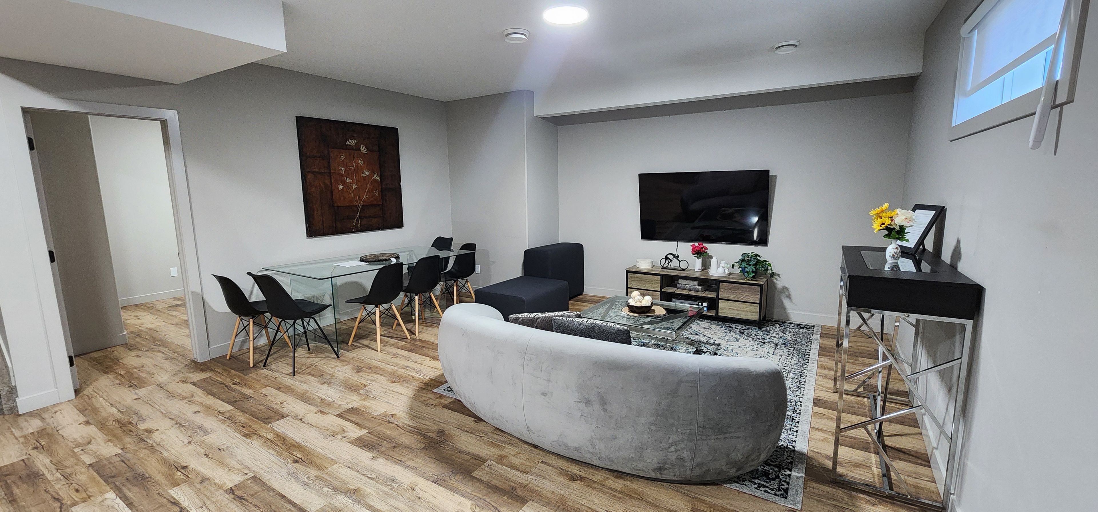 A modern living room with a glass dining table, black chairs, a grey sofa, a coffee table, a flat-screen TV, and a small side table with decorative items.