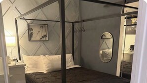 Ein Bett mit Himmel, eine dunkle Bettdecke, ein Nachttisch mit Lampe, ein an der Wand befestigter Spiegel und ein gerahmtes Bild an der Wand.
