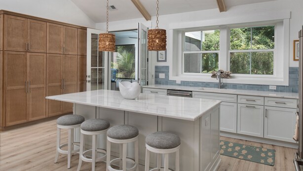 A kitchen with a central island, bar stools, wooden cabinets, and a window with a view of greenery. - Newly built 2bed/2bath house on Dataw Island; TWO WEEK MINIMUM (Saint Helena Island)