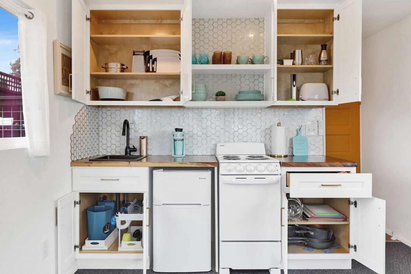 Una cocina compacta con refrigerador, estufa y alacenas abiertas que contienen vajilla y utensilios de cocina.