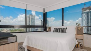 A modern hotel room with a large bed, a desk, and a view of tall buildings through large windows.