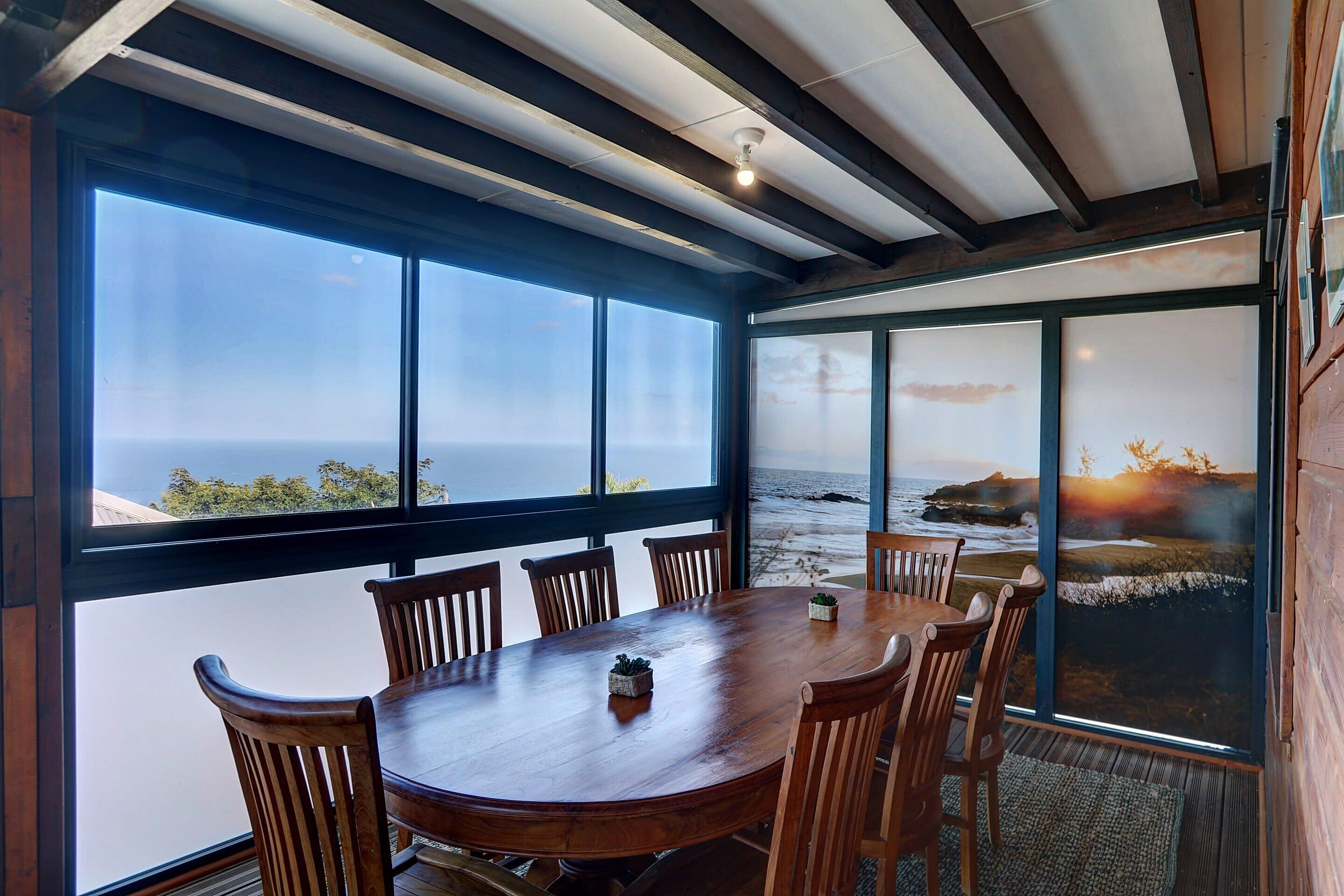 A dining area with a large wooden table and chairs, large windows with ocean views, and a ceiling with exposed beams.