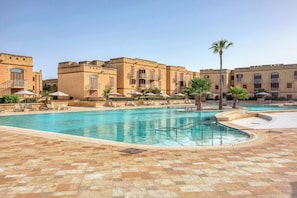 A swimming pool with lounge chairs and umbrellas, surrounded by buildings with balconies. - Fort Chambray Gozo 2BDR Apartment (Ghajnsielem)