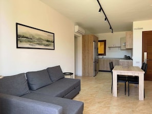 Un saló modern amb un sofà gris, una taula de dinar amb cadires, una zona de cuina amb armaris de fusta i un quadre amb un paisatge al mur.
