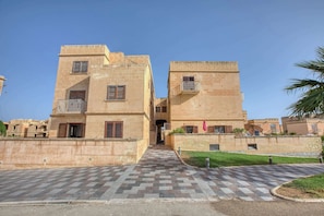 A two-story building with a central entrance, a paved walkway, and a small garden area. - Fort Chambray Gozo 2BDR Apartment (Ghajnsielem)