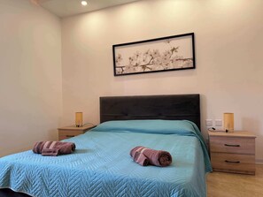 Un dormitori amb un llit, dues taules de nit, un quadre penjat a la paret i una petita taula de nit amb una làmpada.