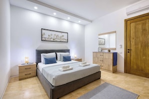 A modern bedroom with a bed, bedside tables, a nightstand, a dresser, a mirror, and a door. - Fort Chambray Gozo 2BDR Apartment (Ghajnsielem)