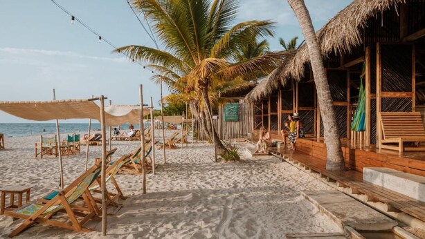 A beach with wooden lounge chairs, a thatched-roof structure, palm trees, and a clear sky. - Dos Aguas Lodge (Rincón del Mar)