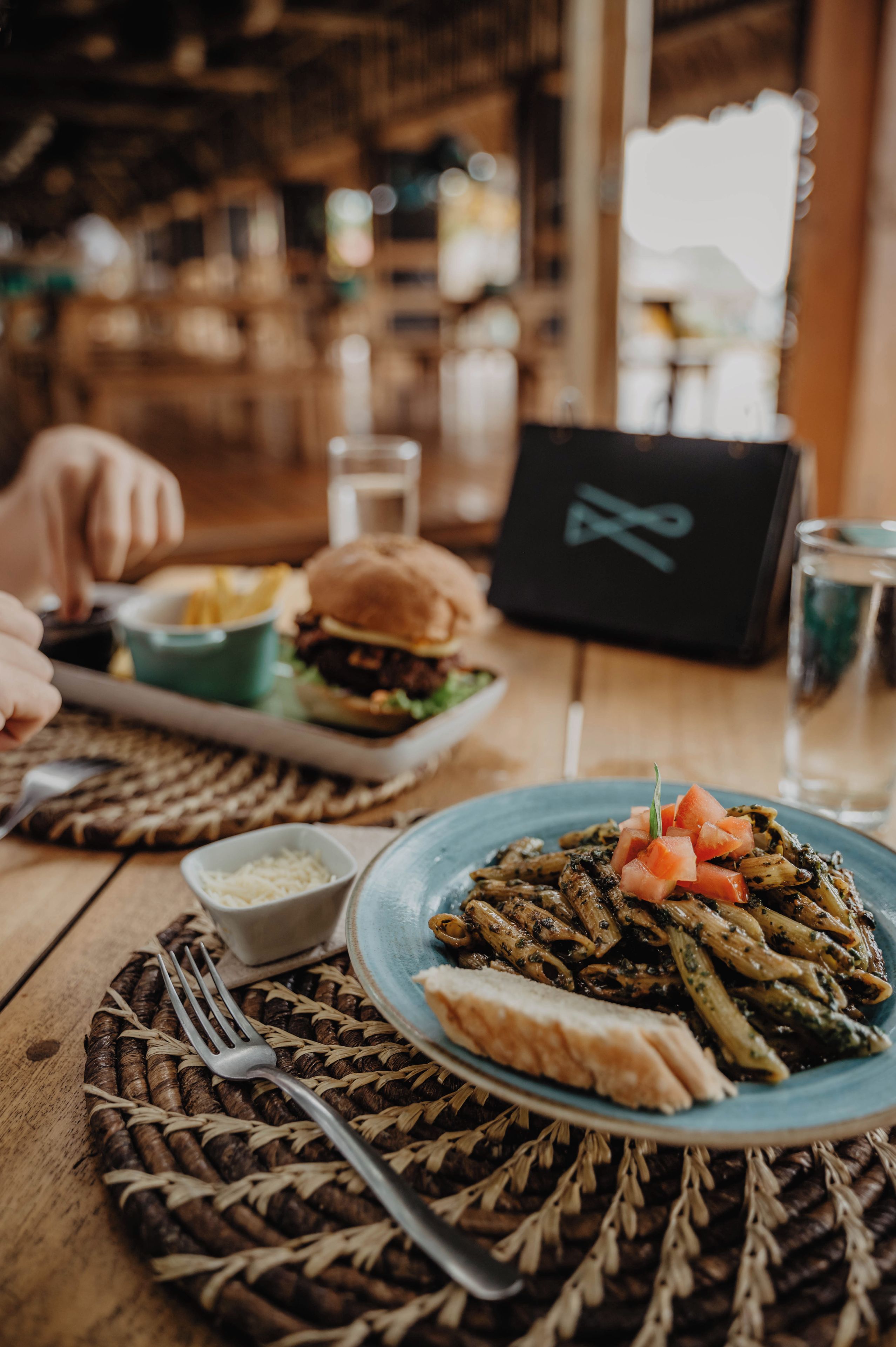 A dining table with a plate of pasta, a burger, and a small bowl of sauce.