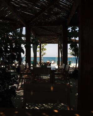 An outdoor dining area with wooden tables and chairs, a thatched roof, and a view of the beach and ocean. - Dos Aguas Lodge (San Onofre)