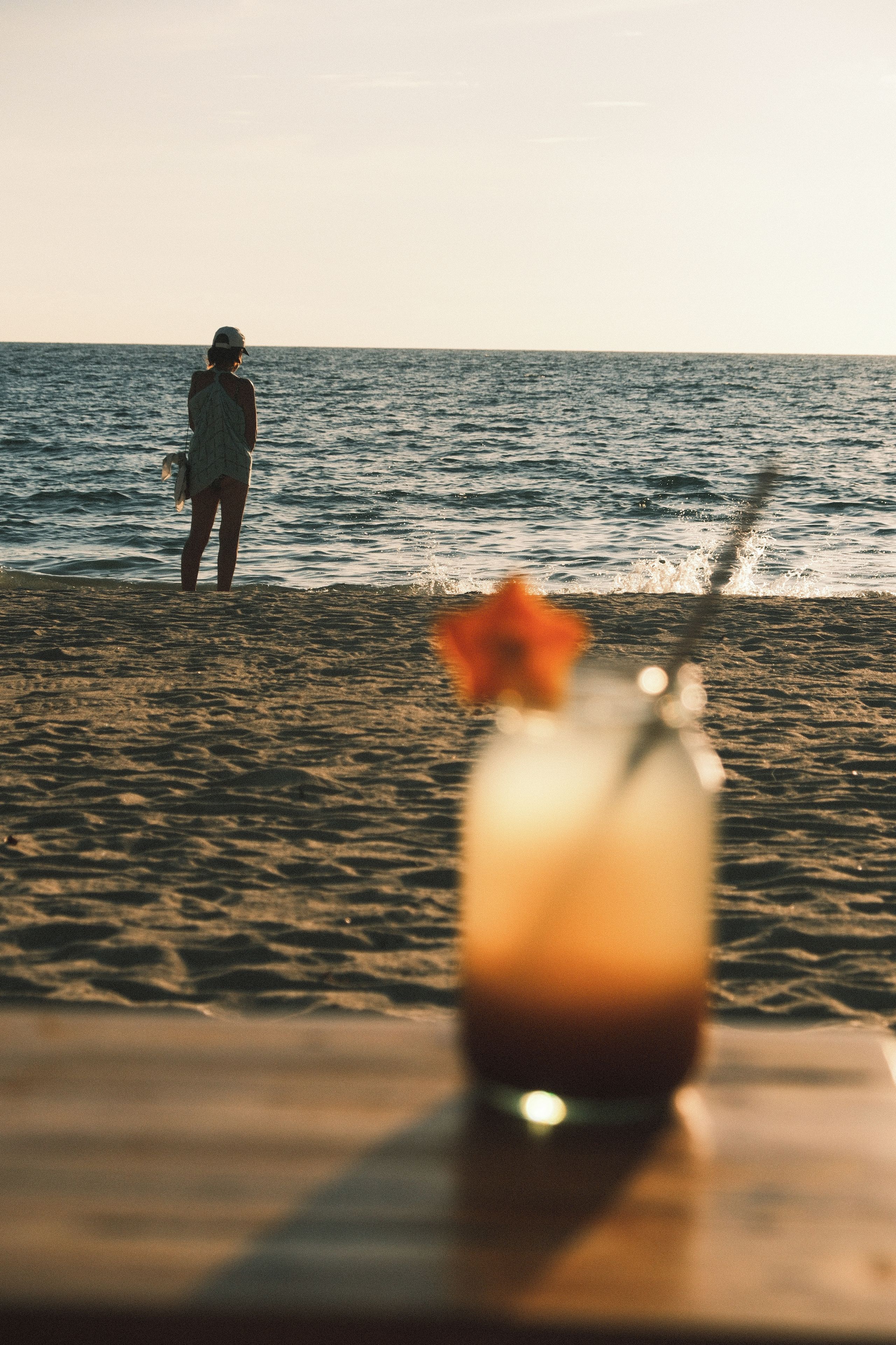 A beach scene with a person standing in the water and a drink on a table in the foreground.