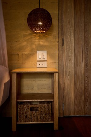 A wooden bedside table with a wall socket and a switch, a woven basket, and a textured pendant light. - Dos Aguas Lodge (San Onofre)