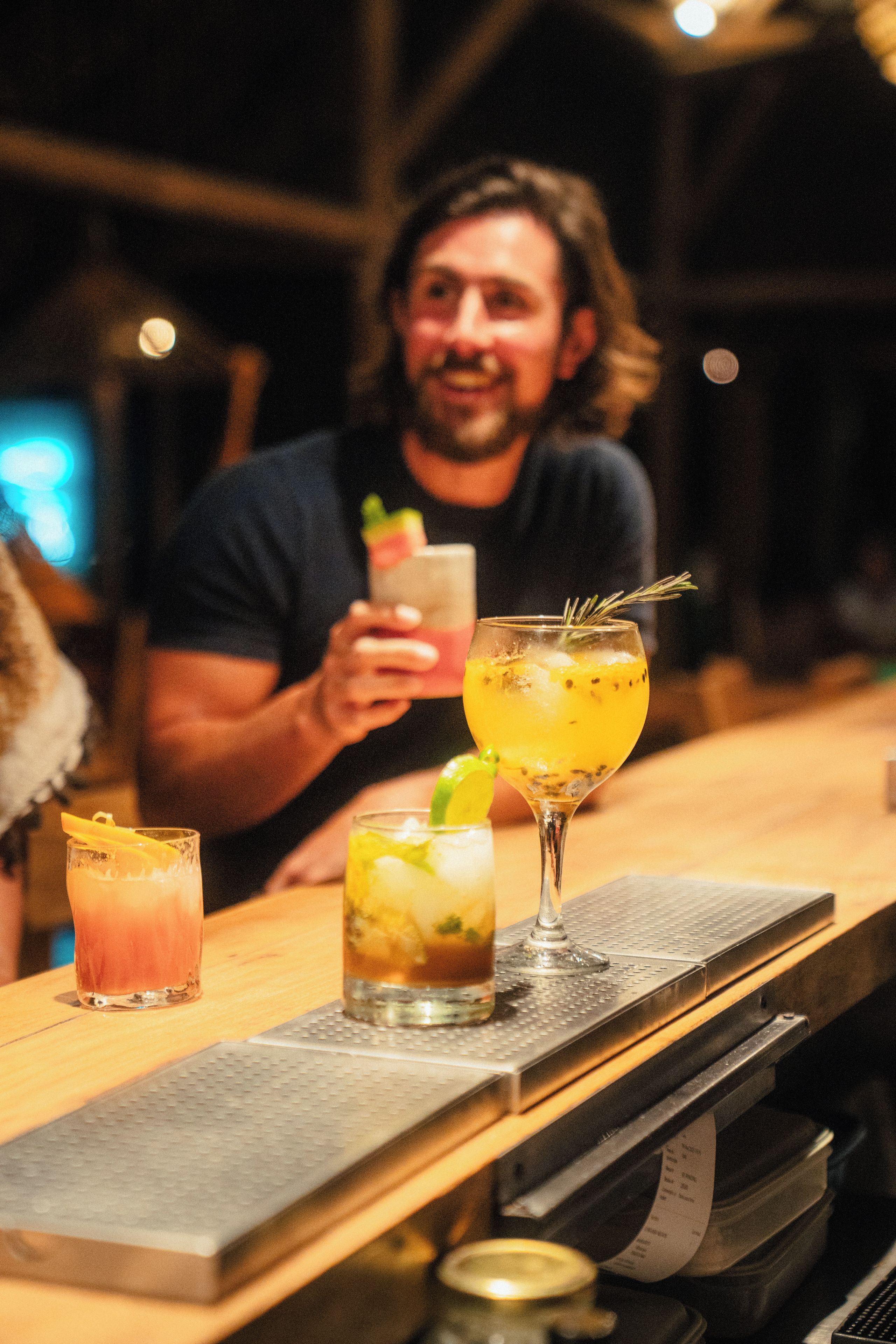 A bar setting with a person holding a drink, a glass with a lemon slice garnish, and a cocktail with ice and a sprig of rosemary.