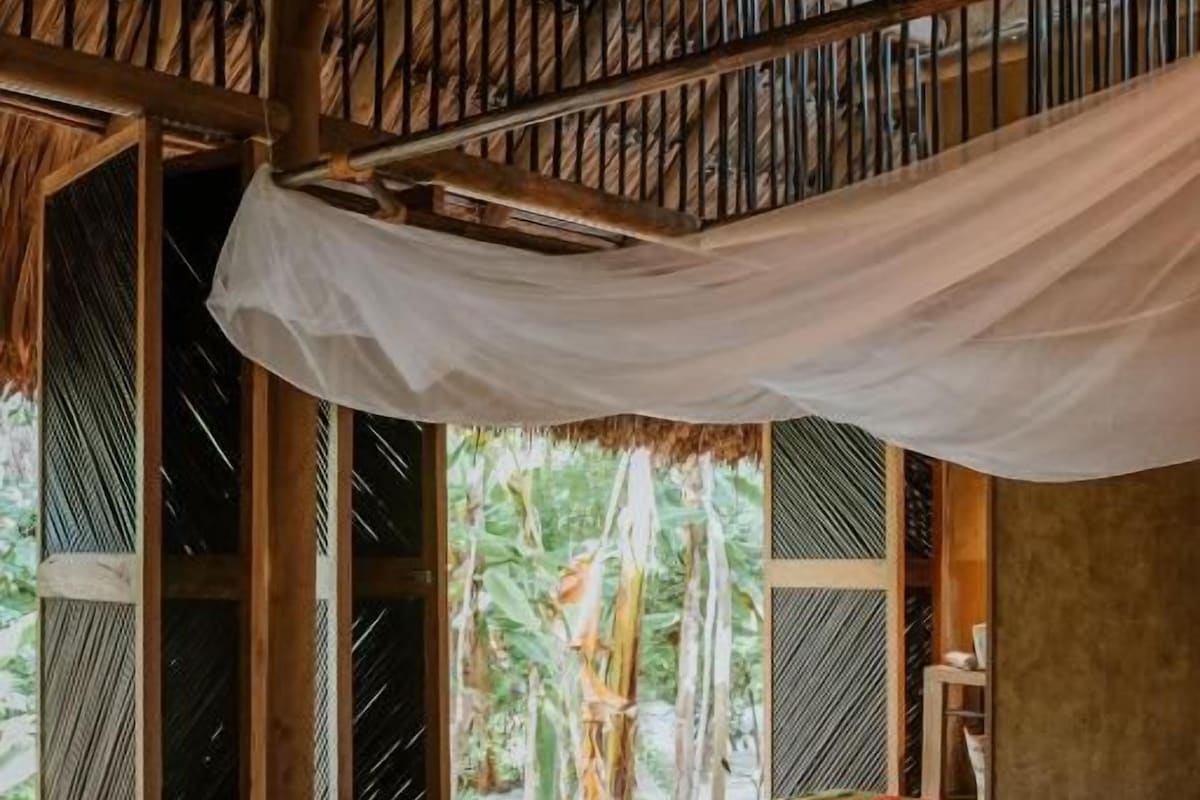 A room with a bed, a window with a view of greenery, and a wooden ceiling structure.