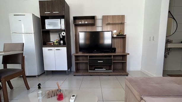 A modern living room with a flat-screen TV, a dining table, a sofa, and a kitchen area with a refrigerator and microwave. - House just 5 minutes from the beach. (Florianópolis)