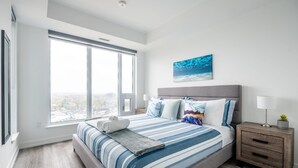 A modern bedroom with a large window, a bed with striped bedding, a nightstand, and a wall-mounted painting.