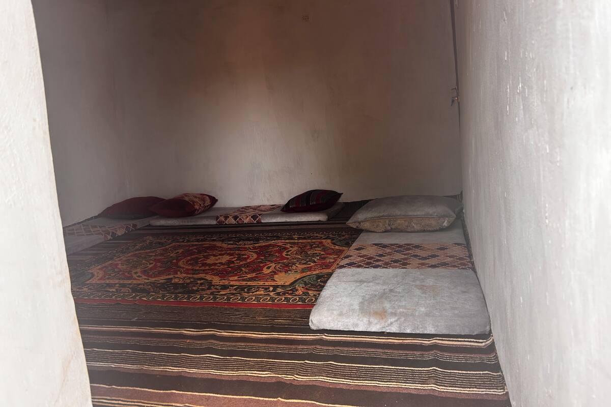 Een kleine, smalle kamer met een bed, kussens en een kamerkleed met patroon.