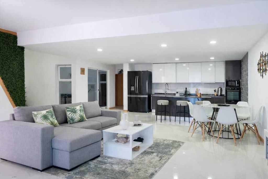 Un salon moderne avec un canapé gris, une table basse blanche et un coin repas avec des chaises blanches.