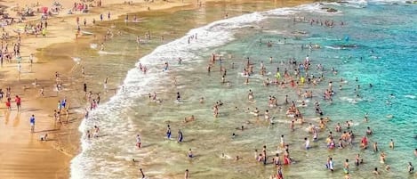 A crowded beach with clear waters and buildings in the background.