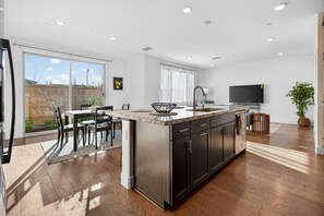 Cocina moderna con una isla grande, comedor con sillas, televisor montado en la pared y puerta corrediza de vidrio que da a un espacio exterior.