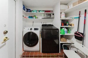 A compact laundry room with a washing machine, dryer, and shelves containing cleaning supplies. - Deluxe Waterfront Villa | BBQ, Pool and Game Room (West Palm Beach)